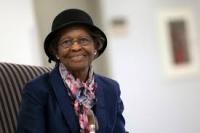 Dr. Gladys West is smiling at the camera, wearing a hat and matching jacket, and a pink/red colored scarf.