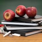 Stack of books with apples on top, chalk and scissors at the base. A chalkboard is in the background.